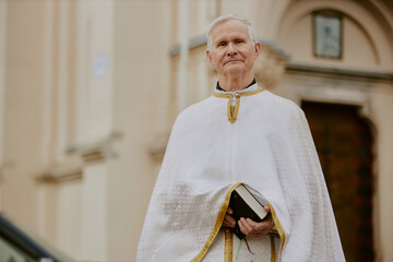 Medium portrait of senior Caucasian Catholic priest holding Bible book standing outdoors looking at...