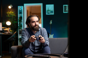 Joyous man playing videogame in home theatre, holding controller. Smiling gamer participating in PvP online multiplayer game using console system and high quality gamepad