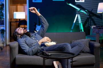 Bored man throwing popcorn in mouth, watching streaming services content on TV, having relaxing evening. Lazy viewer enjoying weekend activity in apartment, munching snacks