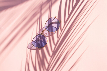 Blue sunglasses on a pink background with contrasting shadows from a palm leaf. View from above