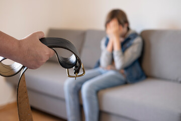 wide leather belt in female parent's hand threatens misbehaving teenage boy sitting on couch,...