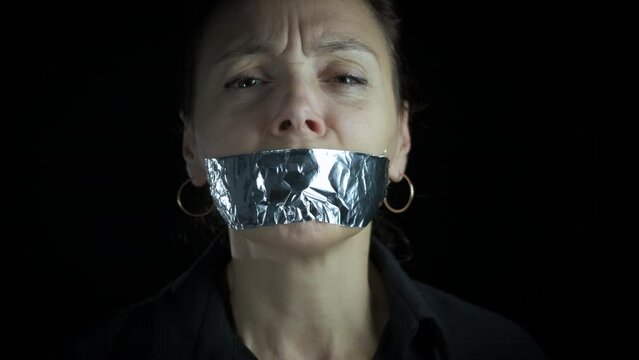 Aggression with mouth on sticky tape. A view of depressed female with closed mouth with tape stand against black background. A concept of home aggression for wife.