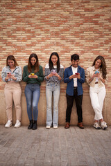 Vertical. Multiracial smiling group young gen z leaning on brick wall using phones outdoors. Cheerful student friends looking at mobile enjoying social media content. Happy device addiction people