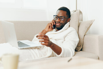 Smiling African American Man Working on Laptop in Home Office, Wearing Bathrobe and Glasses, Talking on Phone