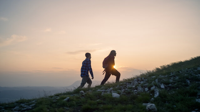 Two teenage hikers walking uphill enjoying a spectacular view while the sun is setting down on the horizon. Active adolescents and nature experience concepts.
