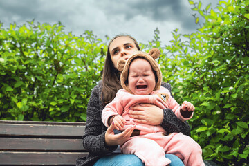 A tired mother sits on a park bench with her baby who is crying hysterically. She looks exhausted...