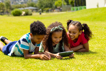 Three biracial children are lying on the grass, looking at a smartphone together