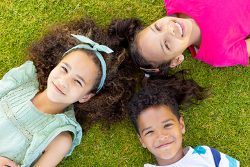 Three biracial children are lying on the grass, smiling up at the camera