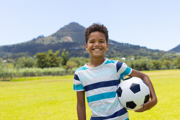 Biracial boy holding a soccer ball, smiling in a sunny outdoor setting