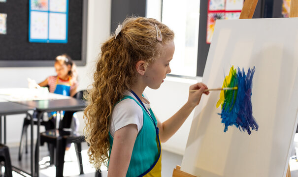 Caucasian girl paints with blue and yellow on an easel in school, biracial girl in the background