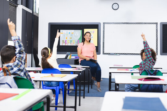 In school, young biracial female teacher smiles at students raising hands in class