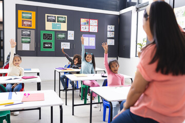 In school, students sitting at desks raising hands, teacher facing them in classroom
