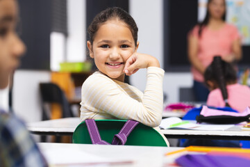 Young biracial girl with brown hair is sitting, smiling at her desk in classroom