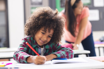 In a school classroom, a biracial young boy writes in a notebook as his mother teaches