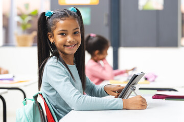 In school, young biracial girl holding tablet smiles at camera in classroom