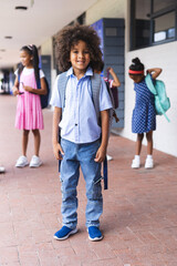In school, young African American boy stands smiling with backpack outdoors