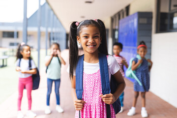 In school, young biracial girl with pink dress and backpack is smiling outdoors