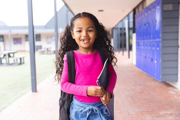 In school, outdoors, young biracial girl with curly hair is holding a notebook