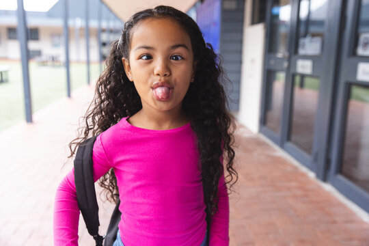 In school outdoors, young biracial girl with a backpack is sticking out her tongue - Powered by Adobe