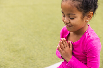 Young biracial girl in pink prays and does yoga on green grass at school with c