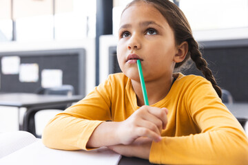 In school, in classroom, young biracial girl holding a pencil, looking thoughtful