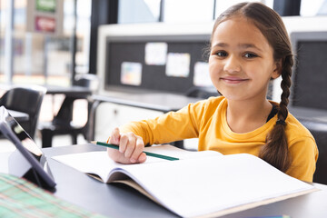 In school, in a classroom, a young biracial girl with light brown skin is writing in a notebook