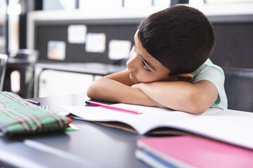 In school, young biracial boy resting head on arms in classroom, looking at notebook