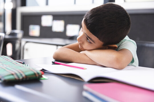 In school, young biracial boy resting head on arms in classroom, looking at notebook - Powered by Adobe