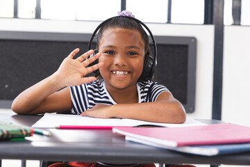 In school, young biracial girl wearing headphones waving at camera in classroom