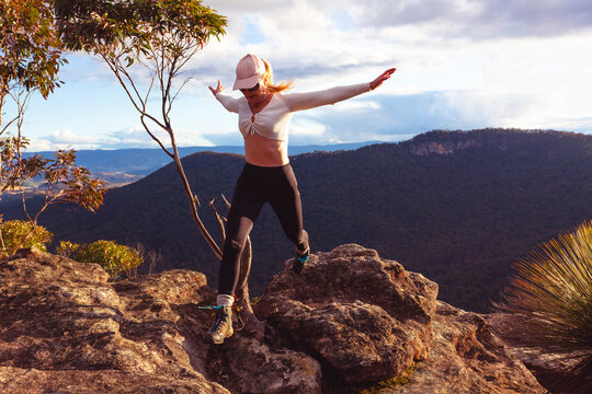 Woman jumping off rocks at cliff ledge with mountain and valley views beyond.