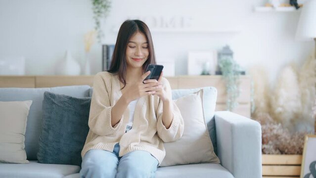 Happy young asian woman relax on comfortable couch at home, typing chat message on smartphone, smiling girl use cell phone chatting, searching information on browser wireless internet, online shopping