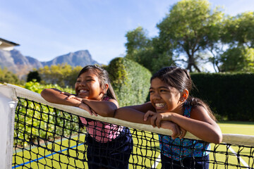Outdoors, two biracial young sisters leaning on tennis net, smiling and having fun