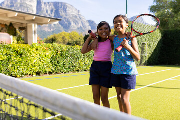 Obraz premium Outdoors, two biracial young sisters smile and hold tennis rackets on tennis court