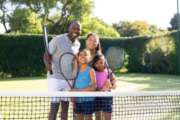 Outdoors, diverse family of four is smiling and holding tennis rackets on sunny day