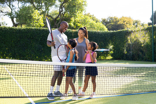 Outdoors, diverse family of four stands on tennis court, holding rackets and smiling