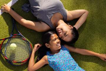 Outdoors biracial mother and daughter lying on grass with tennis rackets, smiling - Powered by Adobe