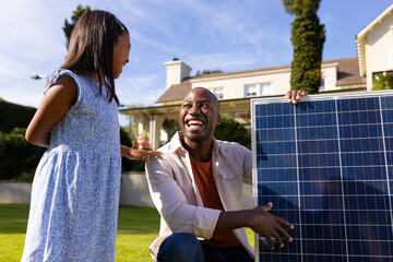 Outdoors, diverse father and daughter smiling at each other holding solar panel