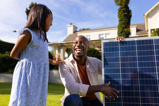 Outdoors, diverse father and daughter smiling at each other holding solar panel