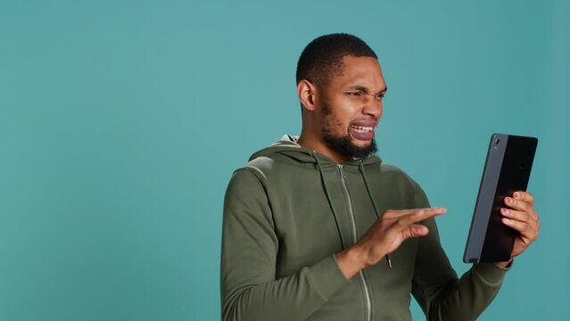 Angry man fighting with friend during videocall on tablet, isolated over studio background. Rude person in disagreement with mate, swearing and pointing finger in teleconference meeting, camera A