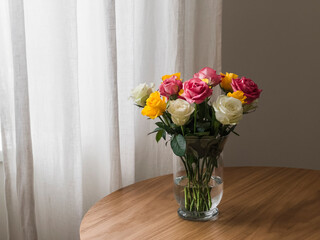 A bright bouquet of yellow, white, pink roses in a glass vase on a wooden table in the living room