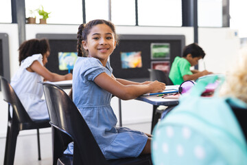 In school, young biracial girl with curly brown hair is sitting, smiling at camera