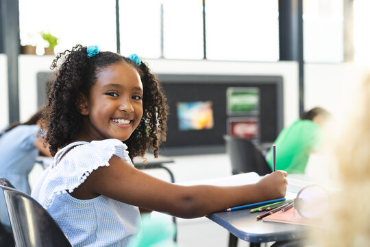 Biracial girl with curly hair smiles while holding a pencil in a school classroom - Powered by Adobe