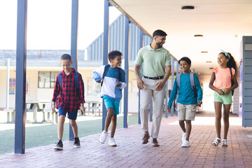 Young Asian male teacher walks with three biracial boys and a biracial girl at school