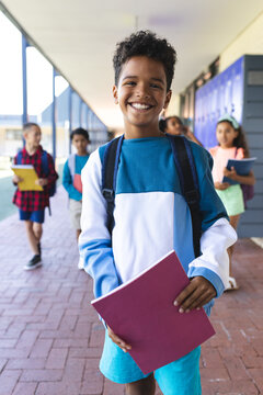 Biracial boy with a bright smile holds a pink folder, standing in a school corridor