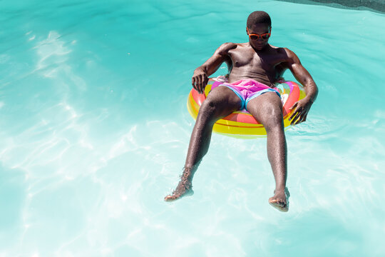 African American man in sunglasses and pink shorts lounges on a pool float outside