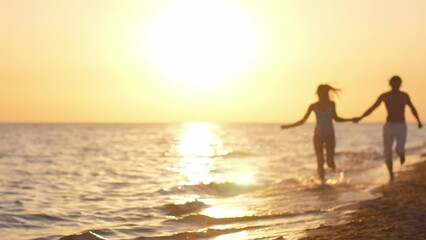 Adorable couple holding hands and running on beach at sunset, silhouetted by bright sun, enjoying evening. A couple runs hand in hand along the beach during sunset, enjoying a joyful and romantic