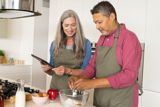At home, diverse senior couple baking in kitchen, Caucasian wife holding tablet