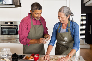 At home, diverse senior couple baking together in kitchen, both smiling in aprons