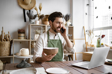 Young bearded man potter taking order for purchase of tableware using phone and laptop. Happy male...