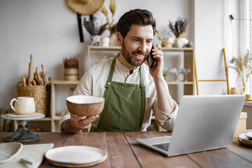 Happy male with new purchase in ceramics shop sitting at table against background of shelves with dishes. Young bearded man potter taking order for purchase of tableware using phone and laptop.